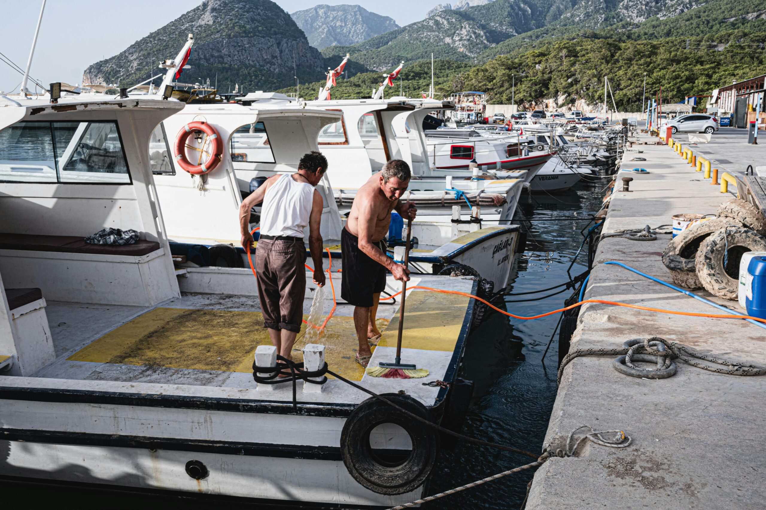 Two men scrubbing boat decks in a marina with mountainous backdrop, enhancing the serene harbor atmosphere.